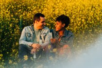 Loving young multiracial couple in casual clothes looking at each other and drinking champagne while sitting in lush blooming meadow during romantic date on sunny day — Stock Photo