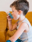 Вид збоку маленького хлопчика з астмою, який використовує інгалятор, сидячи на дивані вдома — стокове фото