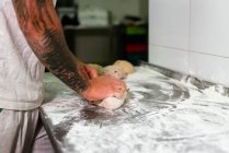 Vista lateral da colheita tatuado padeiro masculino em camisa de pólo branco amassar massa com as mãos, enquanto em pé no balcão de metal na cozinha da padaria moderna — Fotografia de Stock
