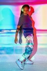 Вид сбоку на модную молодую доминиканскую миллениалку с длинными афро-брейдами, стоящую на полу и смотрящую вниз, слушая музыку в наушниках в комнате с цветной геометрической подсветкой — стоковое фото