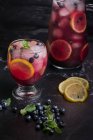 Alto ângulo de vidro e jarro com limonada fria refrescante com mirtilos frescos e fatias de limão colocadas na mesa escura — Fotografia de Stock