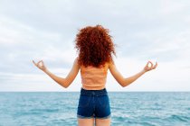 Вид ззаду на невпізнавану жінку з кучерявим рудим волоссям, що робить жест дзен на березі блакитного розривного моря — стокове фото