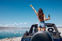 Back view of excited woman in summer outfit raising hand of beer while standing on roof of safari car on shore of river — Stock Photo