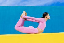 Visão lateral do corpo inteiro fêmea descalça fazendo pose de arco durante o treinamento Dhanurasana pose na parede amarela no fundo azul — Fotografia de Stock