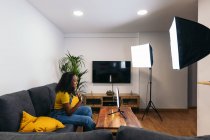 Smiling black female on couch waving hand while using smartphone on LED ring lamp near professional lights on tripods — Fotografia de Stock