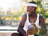 Happy African American male basketball player sitting on sports ground with smartphone on sunny day in summer and looking away — Stock Photo