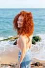 Ottimista giovane femmina con i capelli rossi volanti in piedi guardando la fotocamera sulla costa del mare blu increspato — Foto stock