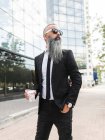 Serious bearded hipster male in formal wear and sunglasses standing on street with takeaway hot drink near modern building in city — Stock Photo