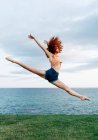 Full body of barefoot female doing split while jumping high with raised arms on coast of rippling sea — Stock Photo
