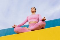 From below of young female in activewear sitting in lotus pose on sports ground while meditating — Stock Photo