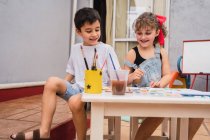 Enfants positifs avec pinceaux peinture avec aquarelles colorées sur papier à table avec des fournitures dans la salle de lumière avec tableau blanc — Photo de stock