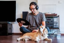 Hombre conectando auriculares al reproductor mientras está sentado en el suelo con Cocker spaniel cachorro en la sala de luz - foto de stock