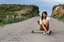 Full body of positive ethnic female browsing on smartphone while sitting on longboard on asphalt road — Stock Photo