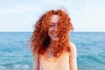 Оптимістична молода жінка з літаючим імбирним волоссям, що стоїть на камеру на узбережжі блакитного розривного моря — стокове фото
