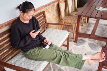 Молодая этническая женщина ласкает милую кошечку во время просмотра мобильного телефона на скамейке — стоковое фото