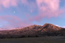 Paysage à couper le souffle d'une chaîne de montagnes rocheuses et d'une vallée avec des arbres verts sous un ciel rose au coucher du soleil avec des nuages et la lune dans le parc national de Sierra de Guadarrama en Espagne — Photo de stock