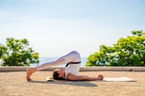 Side view of calm female in white activewear performing Halasana on mat on ground in park against green trees and cloudless blue sky in sunny day — Stock Photo