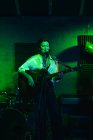 Уверенная дама с гитарой с закрытыми глазами поет в микрофон во время исполнения песни в ярком клубе с неоновым светом — стоковое фото