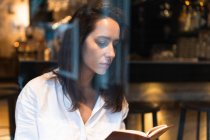 Через окно концентрированной молодой женщины в белой рубашке, читающей книгу, сидя в кафе — стоковое фото