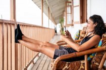 Vista lateral de la alegre joven turista en ropa casual sonriendo mientras está sentada usando un teléfono inteligente en un sillón en una terraza de madera de una casa envejecida en un día soleado - foto de stock