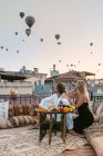 Couple anonyme assis ensemble sur le sol dans un café sur le toit et regardant beaucoup de montgolfières volantes dans la soirée — Photo de stock