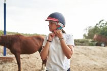 Chica adolescente en ropa casual que se pone el casco mientras está de pie cerca de los caballos con sillas de montar en el patio de la granja en el establo durante el día - foto de stock