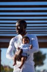 Cuidadoso padre afroamericano en camisa de pie y abrazando al pequeño bebé con el pelo rizado en la calle en un día soleado - foto de stock