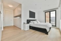 Удобная кровать с белой постельным бельем помещается возле прикроватного столика в просторной спальне с телевизором и стеклянной дверью в стильной квартире — стоковое фото