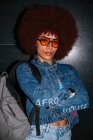 Mulher confiante com penteado afro e roupa na moda e óculos de sol olhando para a câmera enquanto estava de pé com os braços cruzados em fundo preto com mochila à noite — Fotografia de Stock