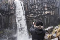 Vista posterior de un viajero masculino irreconocible en ropa interior cálida y sombrero usando un teléfono inteligente mientras toma una foto de la pintoresca cascada de Svartifoss que fluye a través del acantilado de basalto durante el viaje en Islandia - foto de stock