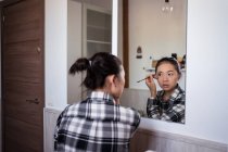 Focused charming ethnic female applying eyebrow pencil while doing makeup and looking in mirror — Stock Photo