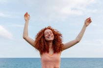 Захоплена жінка з кучерявим довгим імбирним волоссям сміється щасливо, піднімаючи руки і берег рваного моря — стокове фото