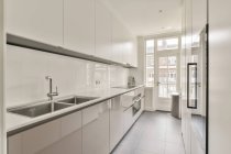 Cuisine étroite ensoleillée avec des meubles blancs de style minimaliste et une porte de balcon dans un appartement contemporain — Photo de stock