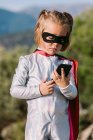 Самоуверенная девушка в костюме супергероя в маске для глаз с накидкой, просматривающей мобильный телефон — стоковое фото