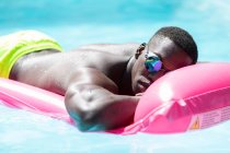 Мужчина без рубашки, афроамериканец в солнечных очках, отдыхает на розовом надувном матрасе в бассейне, загорает в солнечный летний день — стоковое фото