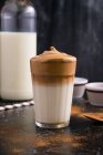 Verre de délicieux café Dalgona avec du lait et garniture mousseuse placée sur une table noire désordonnée avec de la poudre de cacao et du sucre — Photo de stock