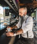 Seitenansicht eines bärtigen Geschäftsmannes mit Tätowierung im edlen Outfit, der Kaffee trinkt, während er in der modernen Cafeteria am Tresen sitzt — Stockfoto