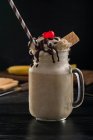 Glass jar of sweet banana split milkshake topped with whipped cream waffles chocolate and cherry on table — Stock Photo