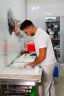 Side view of young tattooed male baker in white polo shirt and eyeglasses kneading dough with hands while standing at metal counter in kitchen of modern bakery — Stock Photo