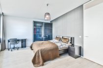 Интерьер современной спальни с подушками и покрытием на кровати напротив стола и шкафа на полу в светлом доме — стоковое фото