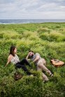 Happy friends musicians with guitar and ukulele sitting on green grass on coast near ocean in nature in daytime — Stock Photo