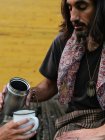 Hippie-Mann gießt Kaffee oder Tee aus einer Thermoskanne in eine Tasse, die eine anonyme Frau auf gelbem Holzgrund hält — Stockfoto