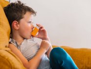 Side view of content sick boy using respirator and breathing oxygen from inhaler while sitting on sofa at home — Stock Photo