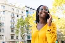 Positive Afroamerikanerin mit Brille schaut weg, während sie auf der Straße mit Wohnhäusern und Bäumen in der Stadt telefoniert — Stockfoto