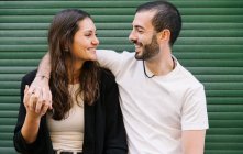Веселая влюбленная молодая латиноамериканская пара в повседневной одежде смеется и смотрит друг на друга, обнимаясь у зеленой стены на городской улице — стоковое фото