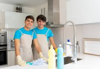Позитивні молоді чоловічі прибиральники в уніформі і гумові рукавички посміхаються і дивляться на камеру під час роботи на сучасній кухні в денний час — стокове фото