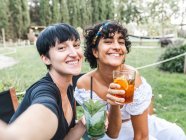 Захоплені багаторасові кращі друзі-жінки в келихах коктейлів, що охолоджуються в літньому парку і приймають селфі на смартфон — стокове фото