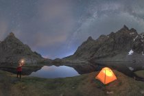 Picturesque scenery of unrecognizable traveler standing with bright light in hand near tent placed among rocky mountains under cloudless night sky milky way located in Circo de Gredos cirque in Spain — Stock Photo