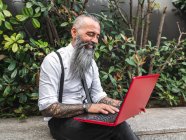 Positive hipster male freelancer in formal wear browsing modern netbook while sitting on stone border near green plants on street during online work — Stock Photo