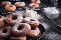 Sweet fried doughnuts served on plate near metal cooling rack and jug of milk on black messy table with powdered sugar — Stock Photo